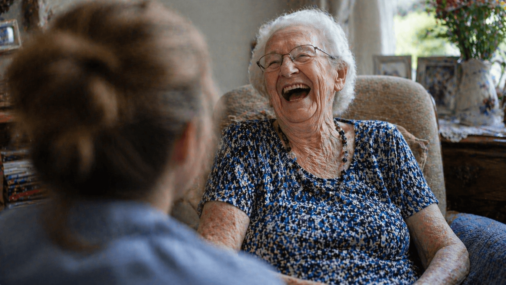 Elderly woman laughing in an armchair while speaking with a younger person, seen from behind, in warm natural light.