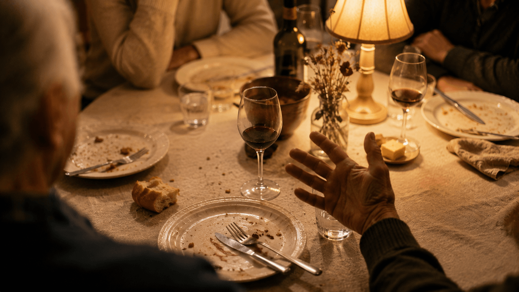 Dinner table after a meal with empty plates, half-full wine glasses, crumbs on a linen tablecloth, and an older hand gesturing mid-conversation in warm lamplight.
