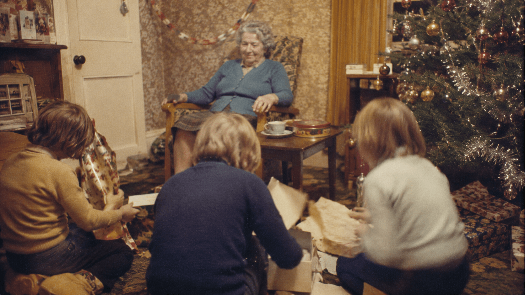 Children opening Christmas presents on the floor while an elderly grandmother watches from an armchair in a 1970s living room.