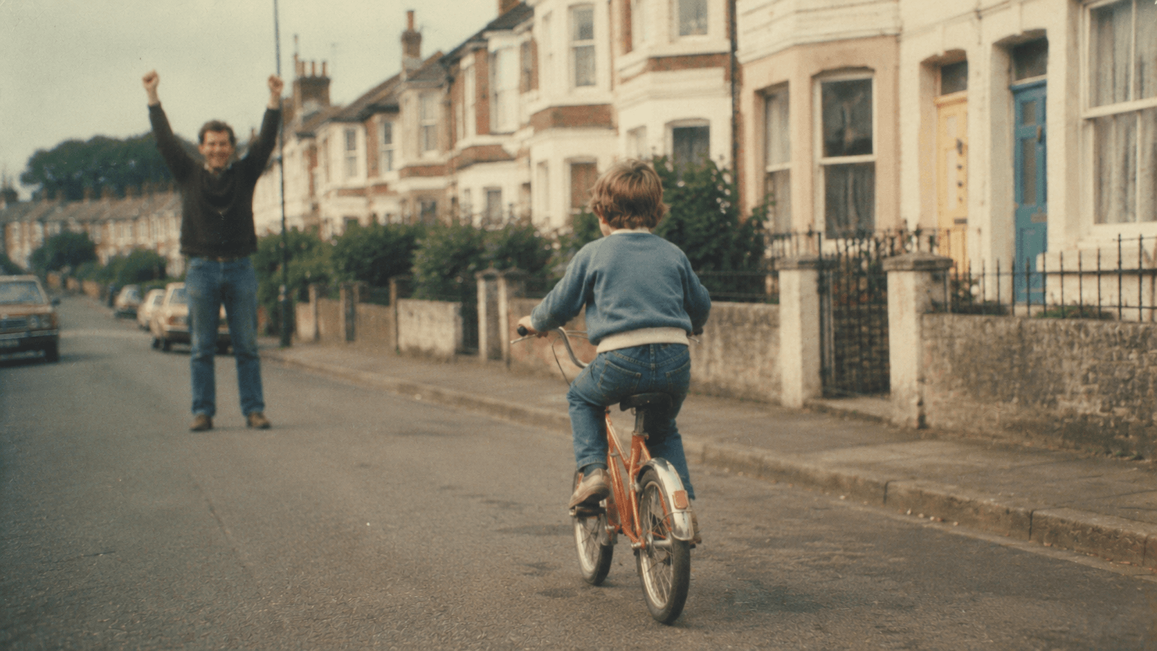 Young child riding an orange bicycle down a suburban street while a father stands behind with arms raised in celebration.